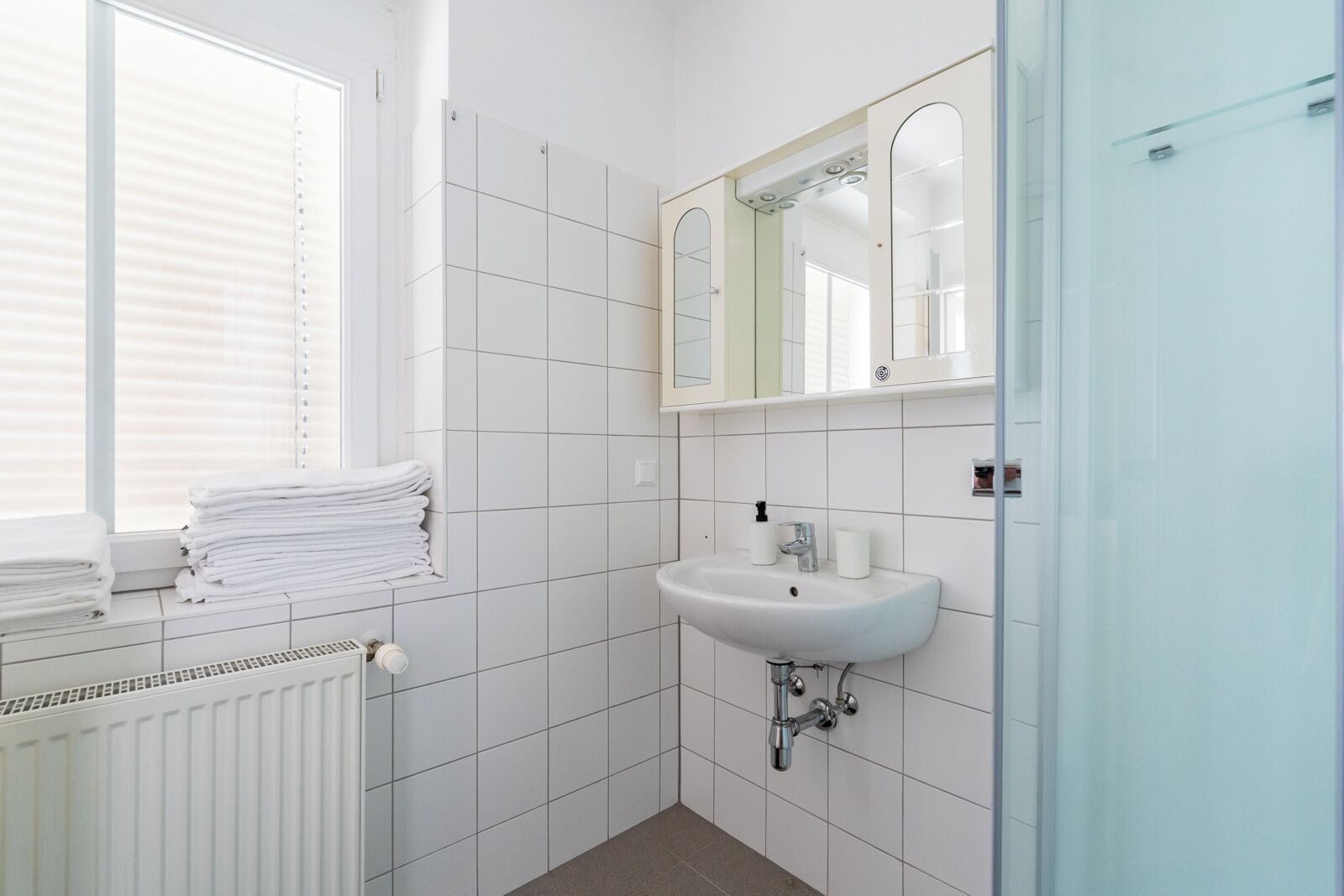 A bathroom with white tiles, a sink, a mirror, and a window with blinds.