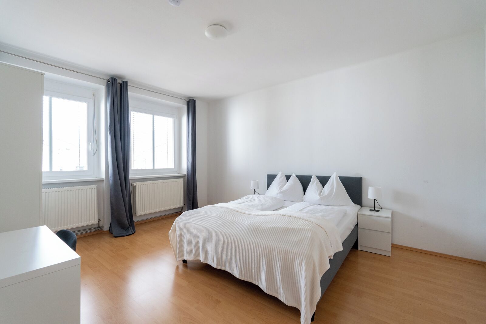A modern bedroom with a bed, bedside lamps, a radiator, and a window with curtains.