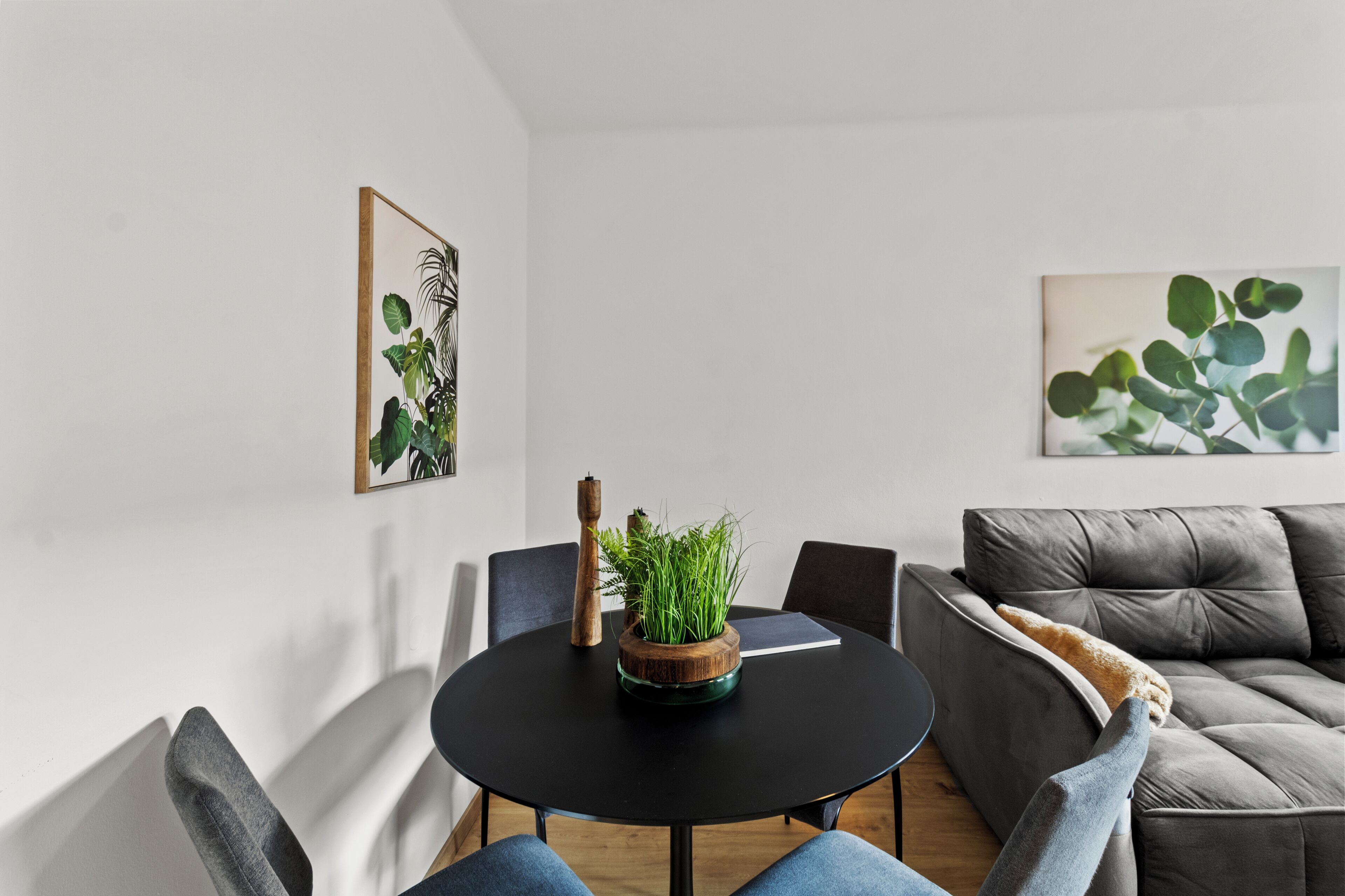 A modern living room with a round black table, a grey sofa, and two armchairs. There are two framed botanical prints on the walls.