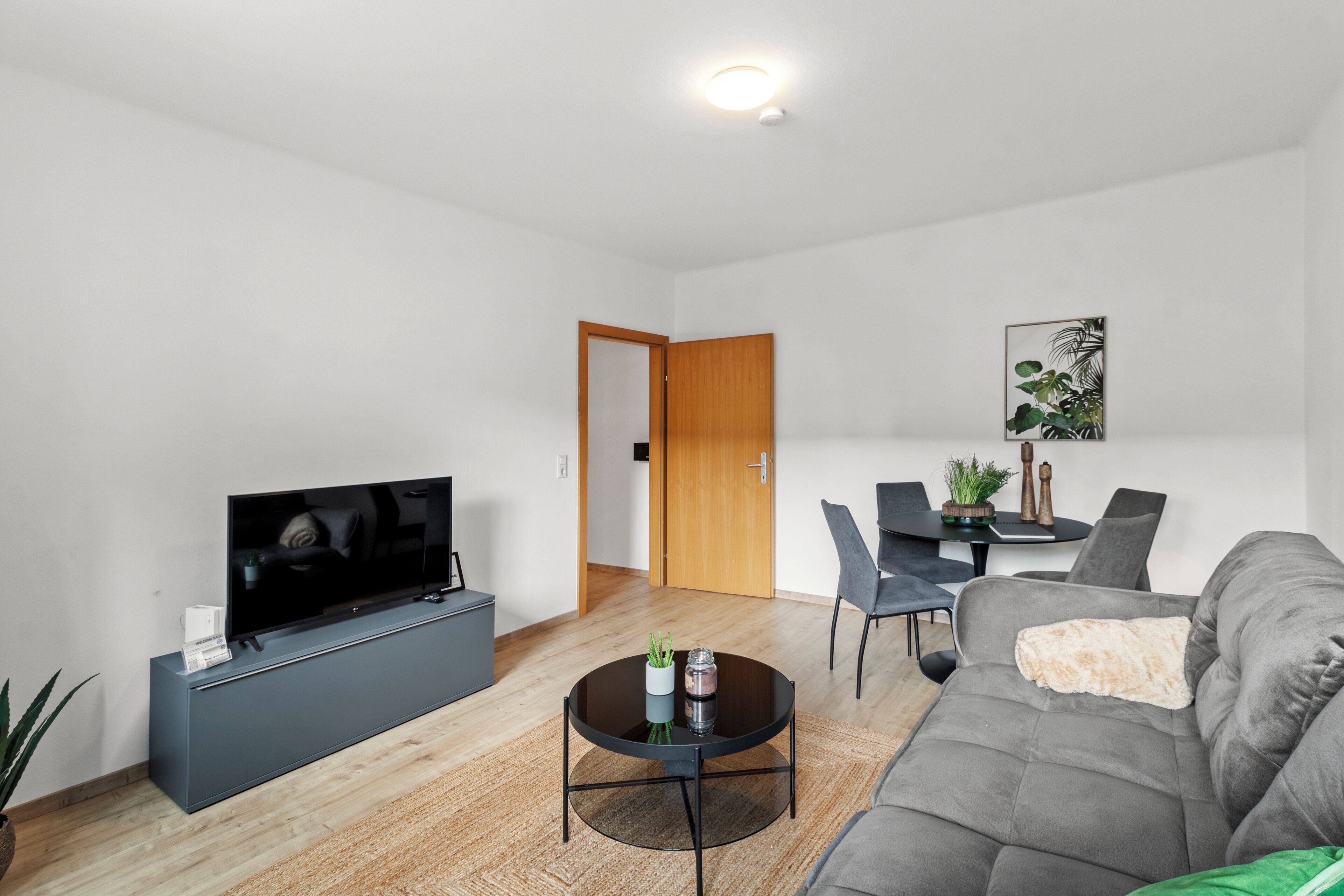 A modern living room with a flat-screen TV, a grey sofa, a round coffee table, and a dining area with chairs and a table.