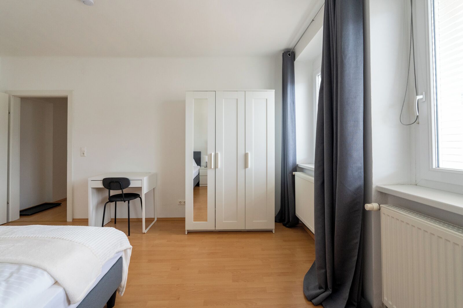 Une chambre à coucher comprenant un lit, un bureau, une chaise, une armoire et une fenêtre avec des rideaux.