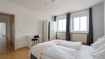 Ein Schlafzimmer mit einem Bett, einem Schreibtisch und einem Sessel. Es gibt zwei Fenster mit Vorhängen.