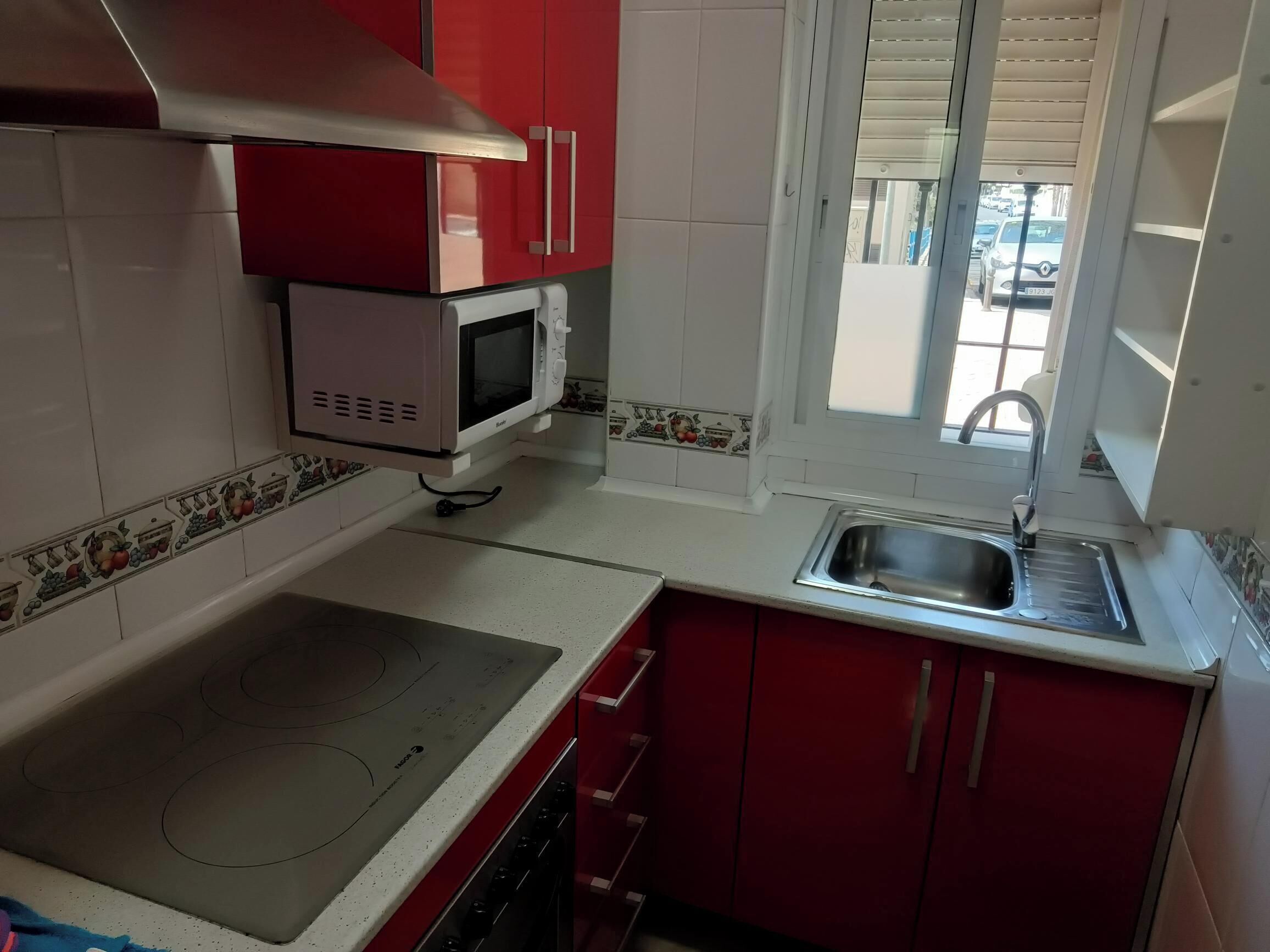 A kitchen with red cabinets, a white microwave, a stainless steel sink, and a window with blinds.