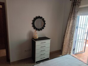 Een slaapkamer met een bed, een dressoir met lades, een spiegel met een zwarte lijst en een fles lotion op het dressoir.