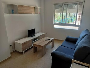 Een woonkamer met een televisie, een salontafel, een bank en een raam met jaloezieën.