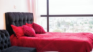 Un lit avec une tête de lit noire et une literie rouge, une petite table ronde et une vue sur le paysage urbain par une grande fenêtre.