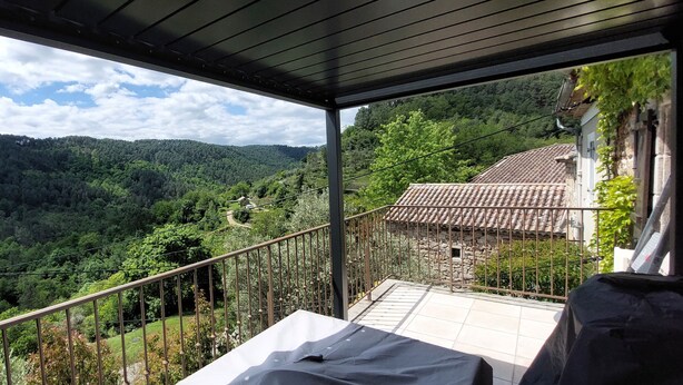 Ein Balkon mit Blick auf ein bewaldetes Tal, ein Gebäude aus Stein mit Ziegeldach und einem Metallgeländer.