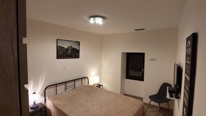 Ein Schlafzimmer mit Bett, Stuhl, Schreibtisch, Lampe, einem Bild an der Wand und einer Tür.