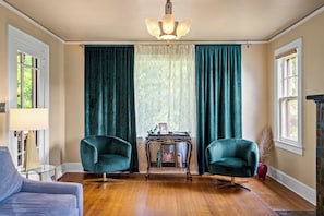 Ein Zimmer mit zwei grünen Sesseln, einem kleinen Tisch mit Büchern, einer Chandelierleuchtre und Blick durch ein mit Vorhängen drapiertes Fenster.