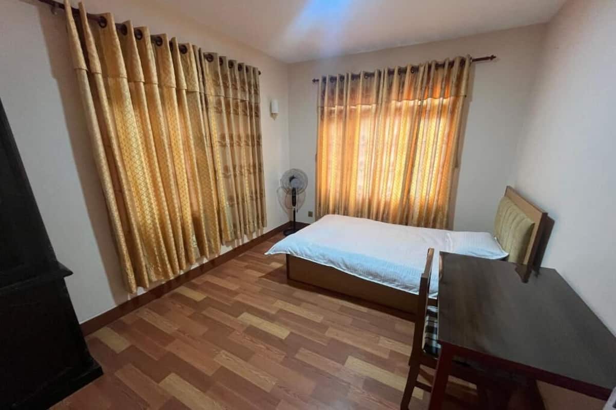 Una habitación con cama, escritorio, silla, ventilador y cortinas.