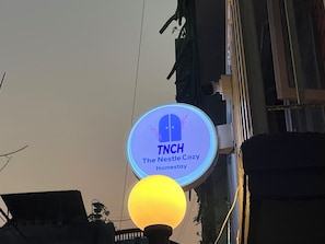 Opastekyltti, jossa lukee ”TNCH The Nestle Cozy Homestay”, ja sen alla on pyöreä logo sekä keltainen, pallonmuotoinen valaisin.