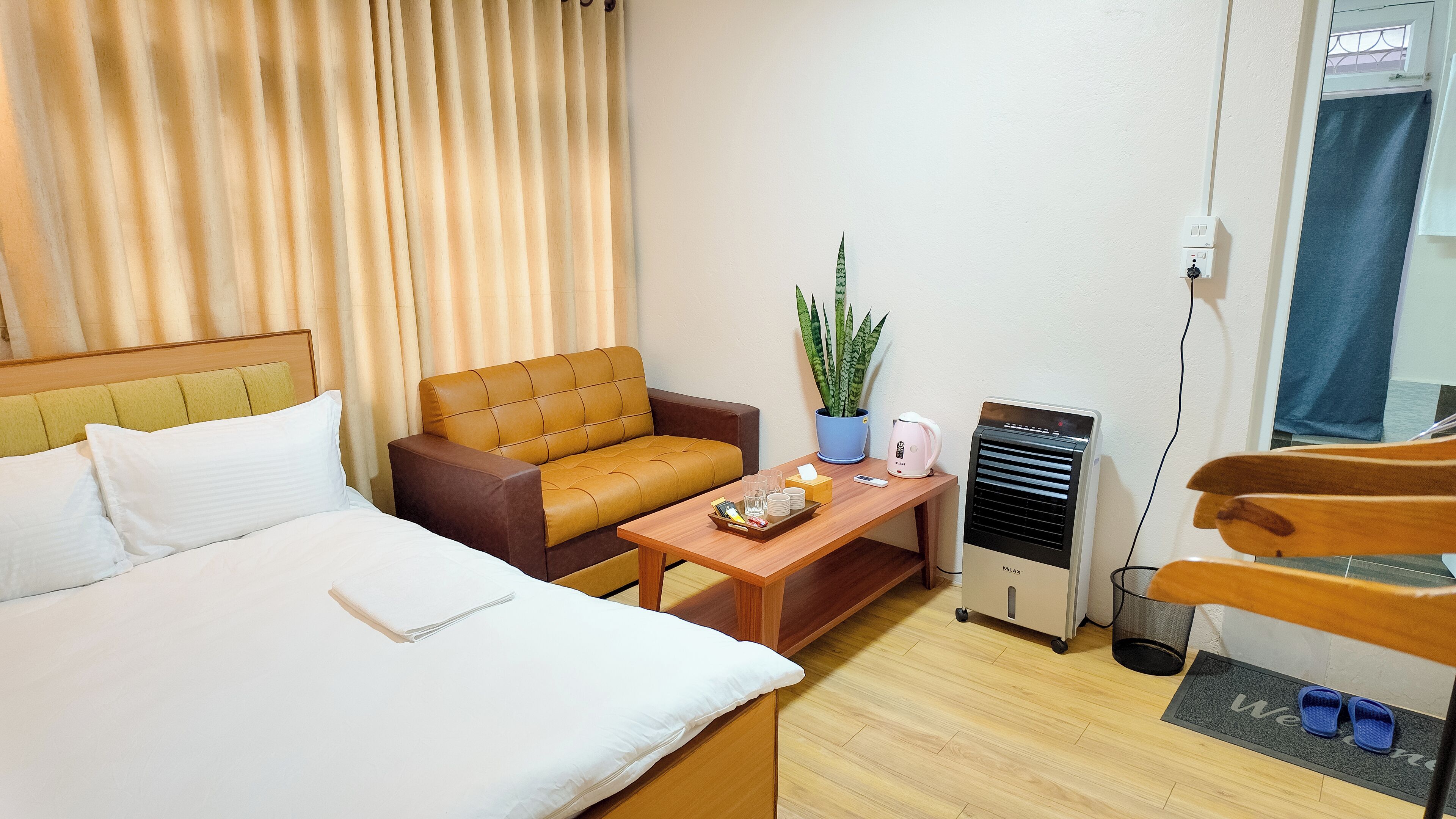 Habitación de hotel con cama, sofá, mesa de centro, planta en maceta, calefactor y un par de pantuflas sobre un felpudo.