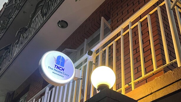 En byggnad med en skylt där det står ”TNCH The Nestle Cozy Homestay” och en tänd lyktstolpe.