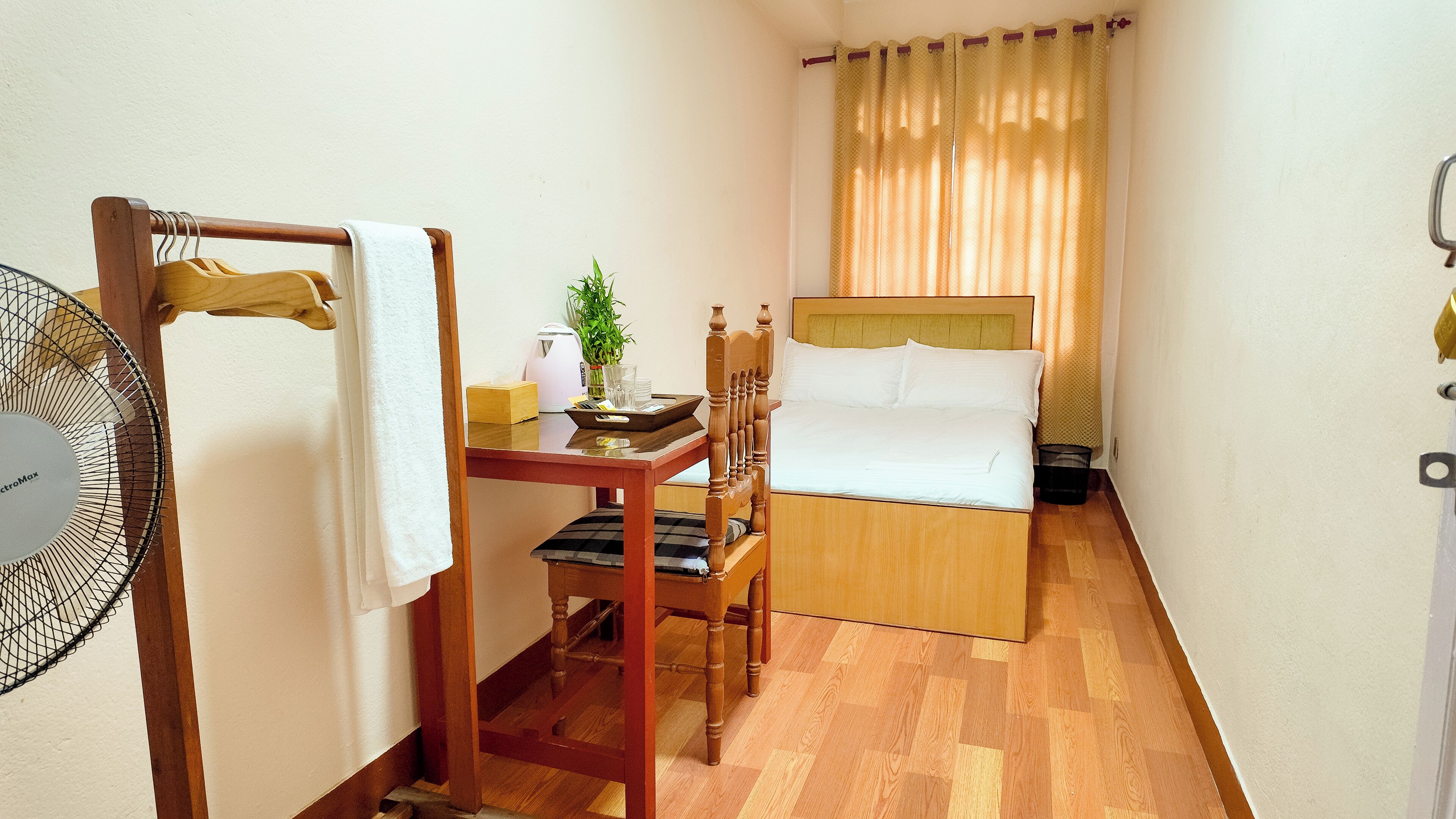 A compact hotel room with a wooden bed, a small table with a chair, a bedside table, a fan, and a window with curtains.