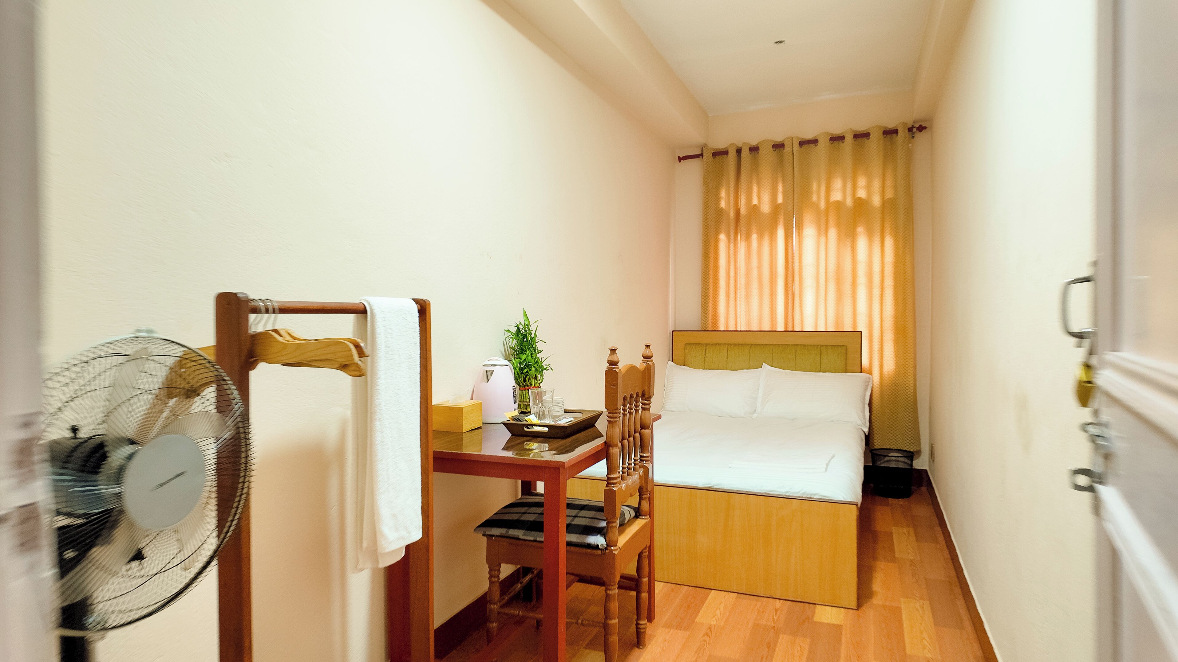 Une chambre d’hôtel de taille réduite, comprenant un lit, une petite table avec une plante, une chaise et un ventilateur sur pied.