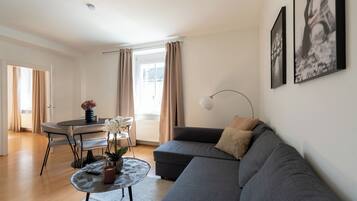Een moderne woonkamer met een grijze bank, een ronde salontafel, een eettafel met stoelen, en een raam met gordijnen.