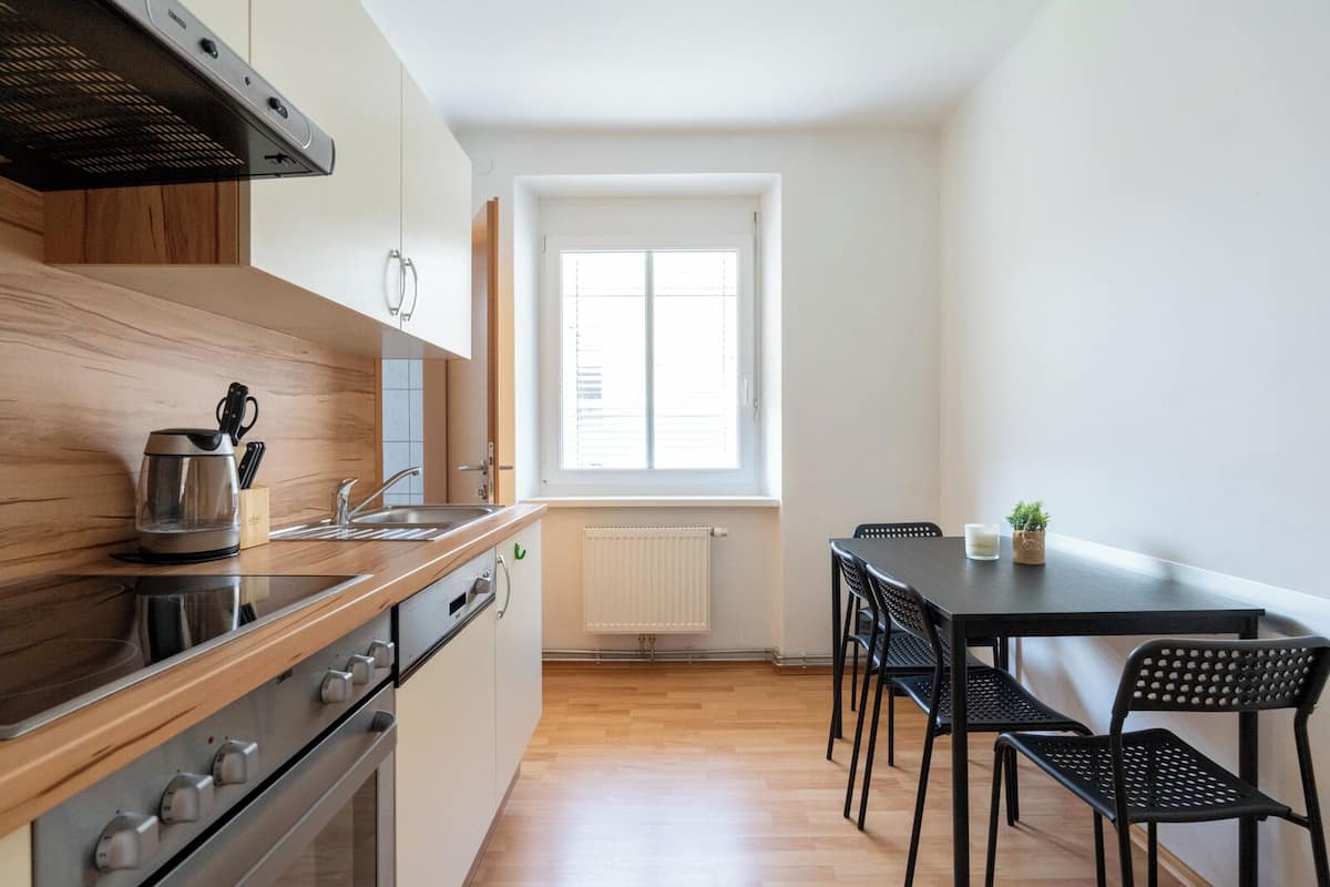 Una cocina moderna con estufa, horno y cafetera, un comedor con mesa y sillas, y una ventana con persianas.