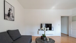 Un salon avec un canapé gris, une table basse ronde ornée d’une plante, un téléviseur posé sur un meuble blanc et un tableau encadré accroché au mur.