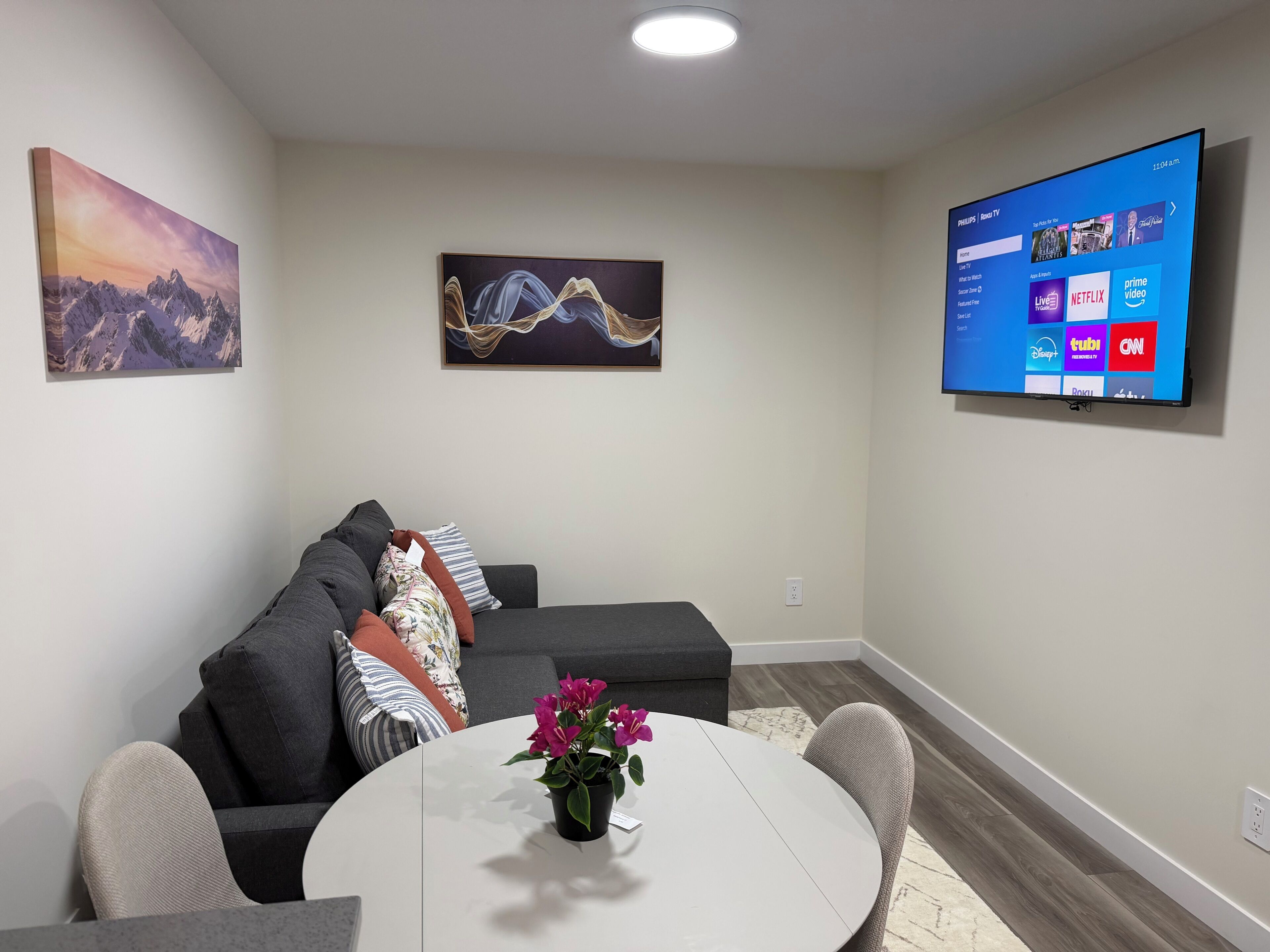 Un salon moderne avec un canapé gris, une table ronde blanche ornée d’un pot de plantes, un téléviseur à écran plat fixé au mur qui affiche l’interface d’un service de diffusion en continu, et deux œuvres d’art encadrées accrochées aux murs.