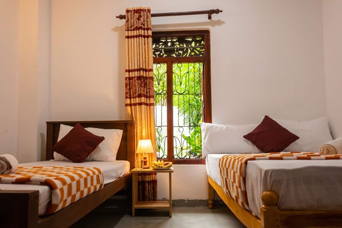 A room with two beds, a nightstand with a lamp, and a window with a view of greenery.