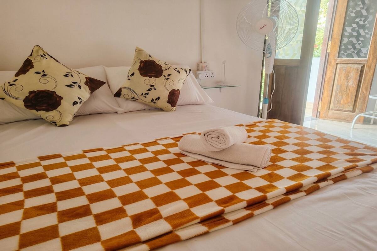 A hotel room with a checkered bedspread, white bedding, floral pillows, a standing fan, and a view of the outdoors.
