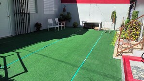 Un patio con césped artificial, sillas blancas, una pequeña mesa y una escalera con detalles rojos.