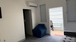 Una habitación con un sofá gris, una silla azul tapizada, un equipo de aire acondicionado blanco, una puerta que da a otra habitación y una ventana con persianas.