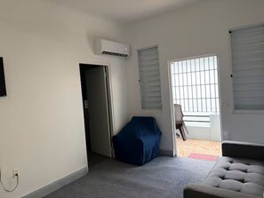 A room with a grey sofa, a covered blue chair, a white air conditioning unit, a door leading to another room, and a window with blinds. - Central & Modern Apt in Miramar (San Juan)