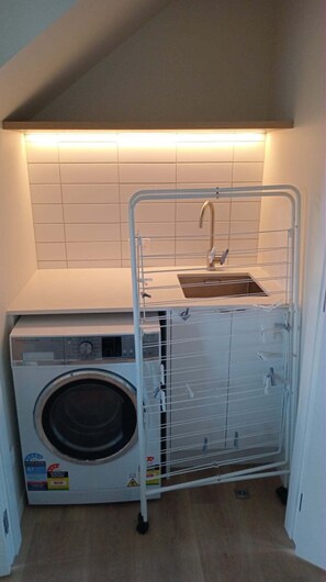 A compact laundry area with a washing machine, a metal drying rack, and a sink under a sloped ceiling.