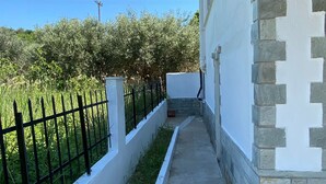 Un estrecho sendero de concreto que lleva a un edificio con fachada de piedra y blanco, delimitado por una cerca de metal negro y follaje.