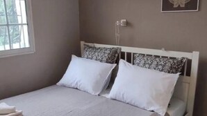 Una cama bien hecha con cabecera blanca, una ventana con persianas y una lámpara fijada a la pared.