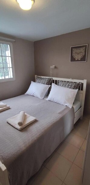 A neatly made bed with a white headboard, a window with blinds, and a wall-mounted light fixture.