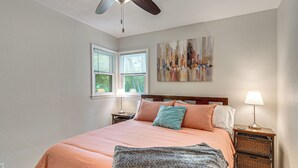 Un dormitorio con cama, ventilador de techo, ventana con persianas, un cuadro en la pared, lámpara de noche y mesita de noche con una lámpara y una cesta.