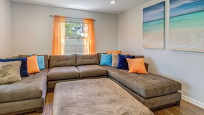 Una sala de estar con un sofá modular, una ventana con cortinas naranjas y dos cuadros con motivos marinos en la pared.