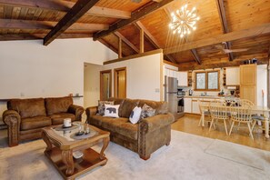 A spacious living area with a brown leather sofa, a wooden coffee table, and a dining table with chairs. A kitchen is visible in the background with wooden cabinets and a refrigerator. - Waterfront Deck + Views: Schroon Lake Sanctuary (Schroon Lake)
