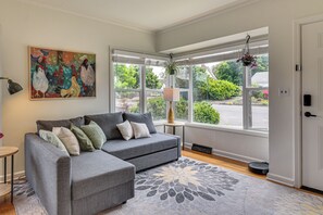 Ein modernes Wohnzimmer mit einem grauen Ecksofa, einem großen Fenster mit Blick ins Grüne und einem dekorativen Teppich.
