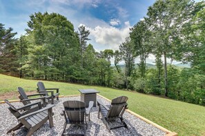 An outdoor seating area with Adirondack chairs and a table, surrounded by a gravel patio and a grassy lawn, with a forested backdrop. - Fire Pit w/ Mtn Views! Blue Ridge Cabin Retreat (Blue Ridge)