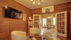 A wooden-paneled room with a flat-screen TV, white armchairs, a coffee table, and a chandelier. - Pet-Friendly Historic Home: Downtown Breaux Bridge (Breaux Bridge)