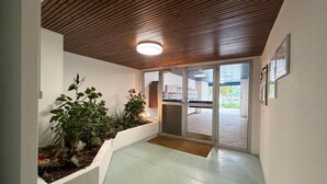 Een gang met een houten plafond, plantenpotten en een glazen deur naar buiten.