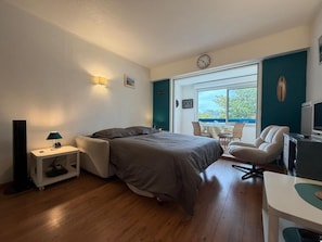 A bedroom with a bed, bedside table, lamp, chair, desk, and a window with a view. - La Brise Marine - Downtown - Port view (CAP BRETON)