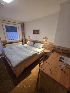 A bedroom with a wooden bed, a small wooden table, a window with a curtain, and a wall-mounted light fixture. - Waldlandhaus (Goslar)
