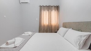 Hotellihuone, jossa on sänky, sängynpääty, ikkuna verhoilla ja seinälle asennettu ilmastointilaite.
