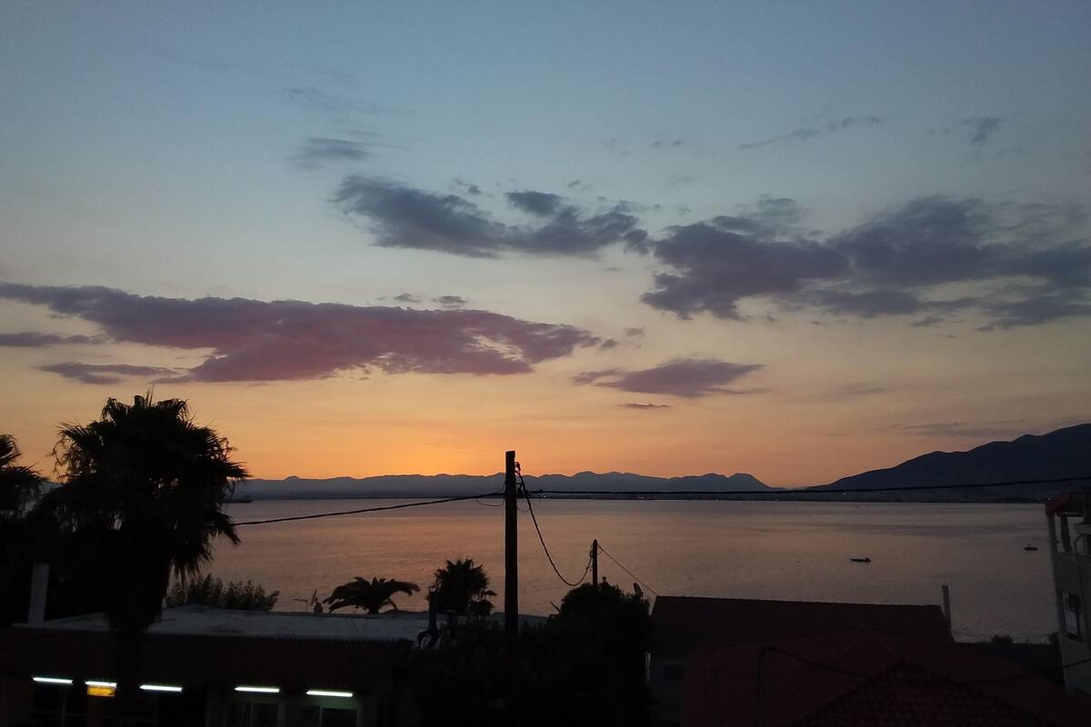Un atardecer sobre una masa de agua tranquila con montañas lejanas, palmeras y un edificio con techo de tejas.