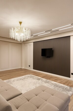 En moderne stue med en lysekrone, et fladskærms-tv monteret på en væg af træpaneler og en beige sofa.