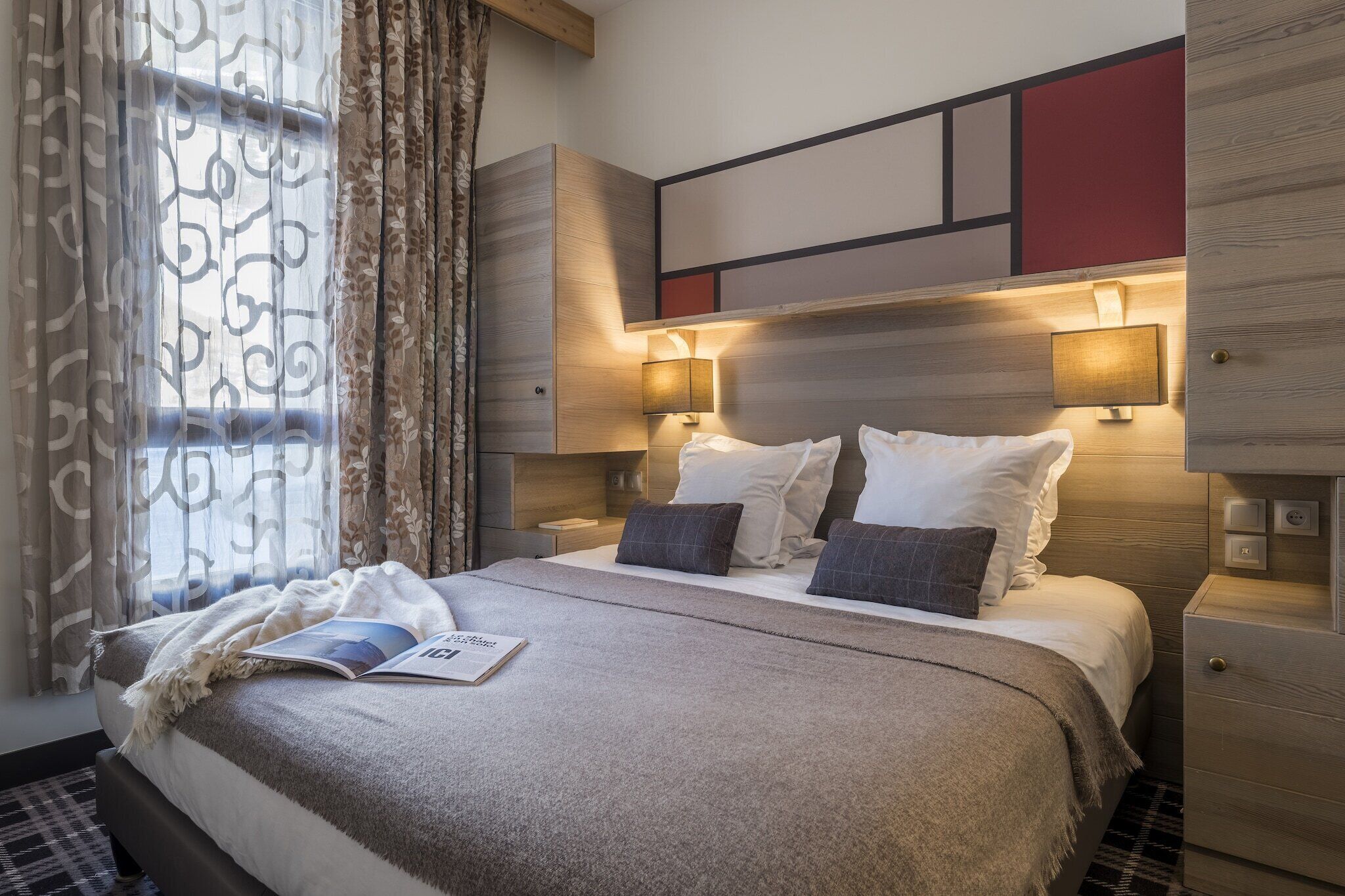 Ein Hotelzimmer mit Bett, Nachttischlampen, einem Fenster mit Vorhängen und einer Zeitschrift auf dem Bett.