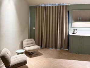 A room with a herringbone floor, a cushioned armchair, a small round table, a window with a curtain, and a kitchenette area. - Romantic Getaway in Alicante (Alicante)
