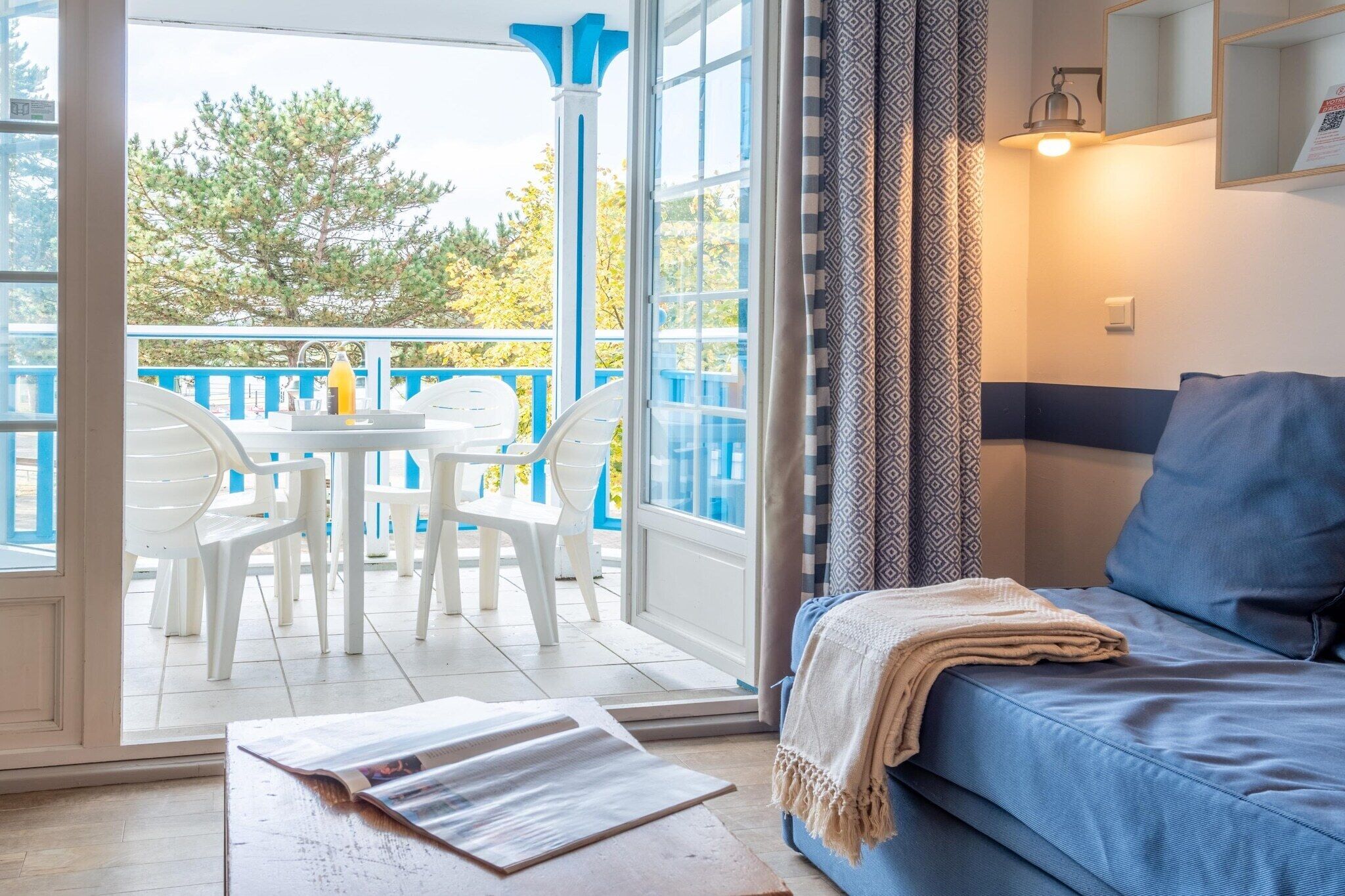 A room with a blue sofa, a small table with a magazine, and a balcony with white chairs and a bottle.