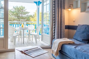 Une pièce avec un canapé bleu, une petite table sur laquelle repose un magazine, et un balcon avec des chaises blanches et une bouteille.