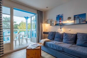 Une pièce avec un canapé bleu, une table en bois, un balcon avec des chaises blanches et des œuvres d’art accrochées aux murs.