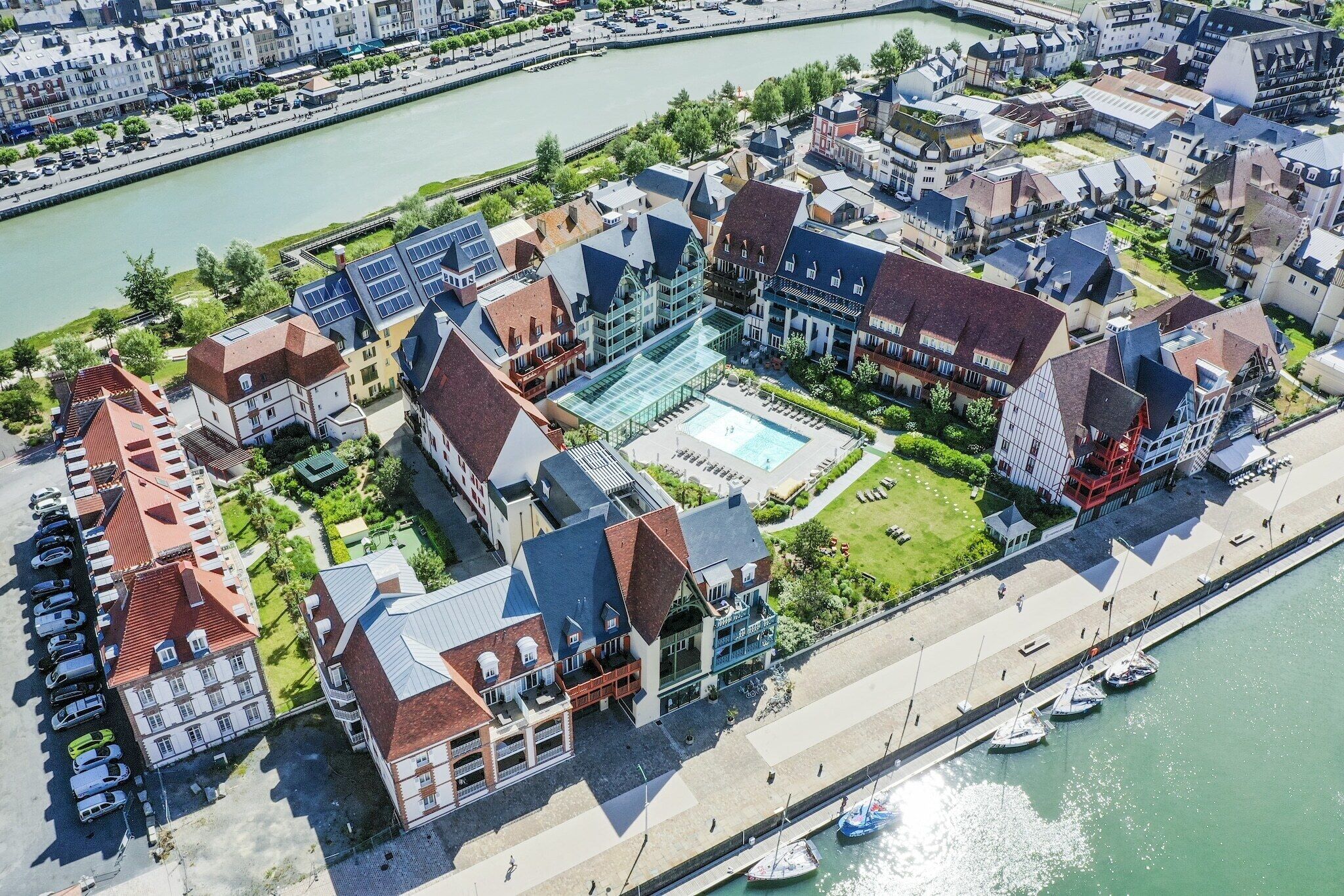 An aerial view of a riverside hotel with a swimming pool, boats, and a promenade.